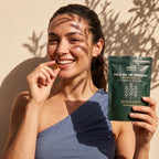 Smiling woman holding a Nura Wild Oil of Oregano softgel and pouch in natural sunlight