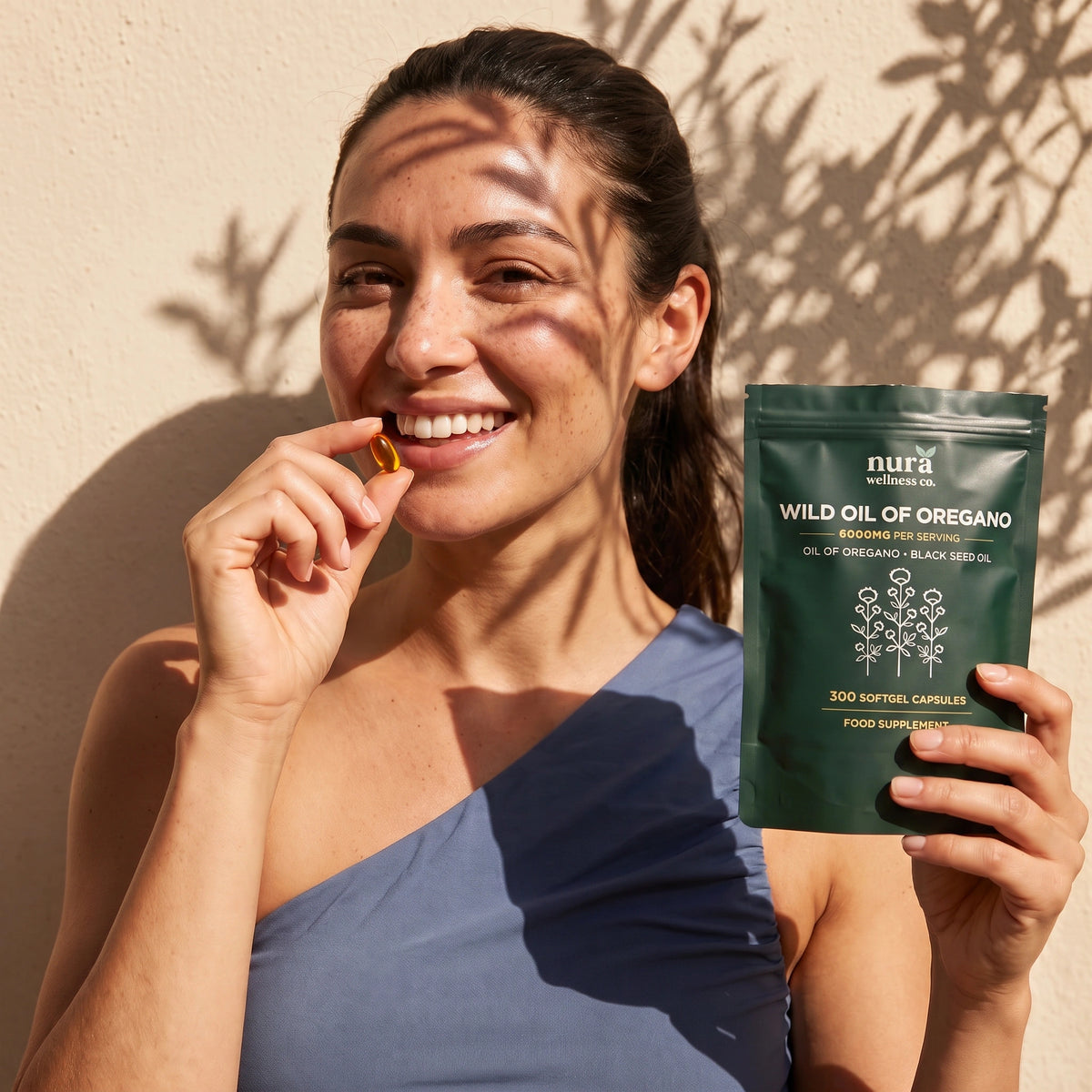 Smiling woman holding a Nura Wild Oil of Oregano softgel and pouch in natural sunlight
